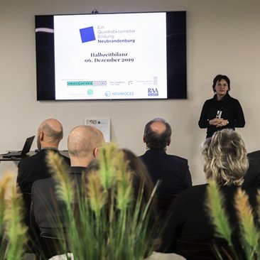 Eine Frau steht vor einer Gruppe neben ihr wird eine Präsentation abgespielt