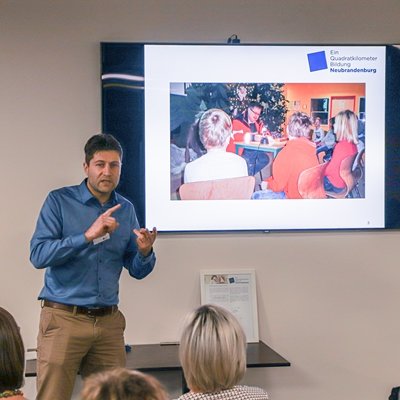 Ein Mann präsentiert etwas vor einer Gruppe, im Hintergrund wird ein Foto gezeigt