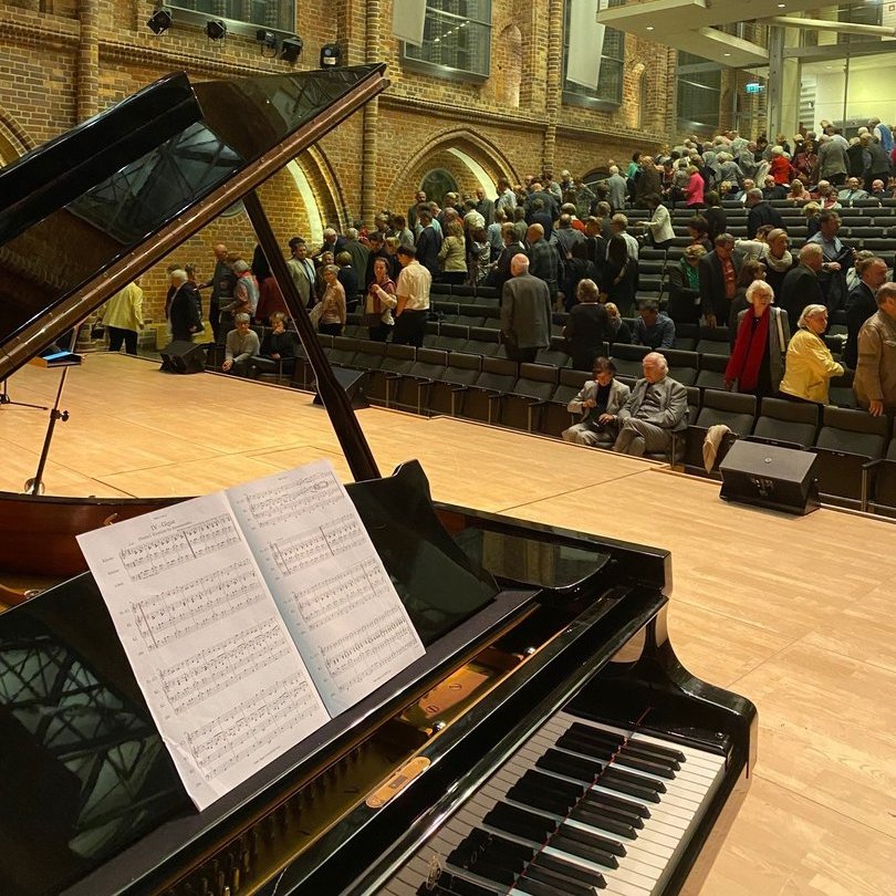 im Vordergrund steht ein Klavier, dahinter nehmen Menschen in Sitzreihen gerade platz 